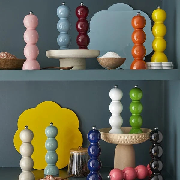 Colourful salt and pepper mills in pink, blue, red, orange, yellow, green, white and black, decoratively arranged on blue-grey shelves with stylish home accessories.