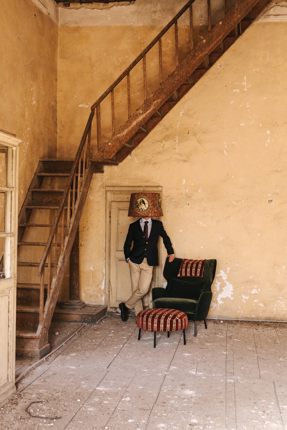 Green velvet armchair with a patterned footstool and a man wearing a lampshade on his head in an old stairwell.