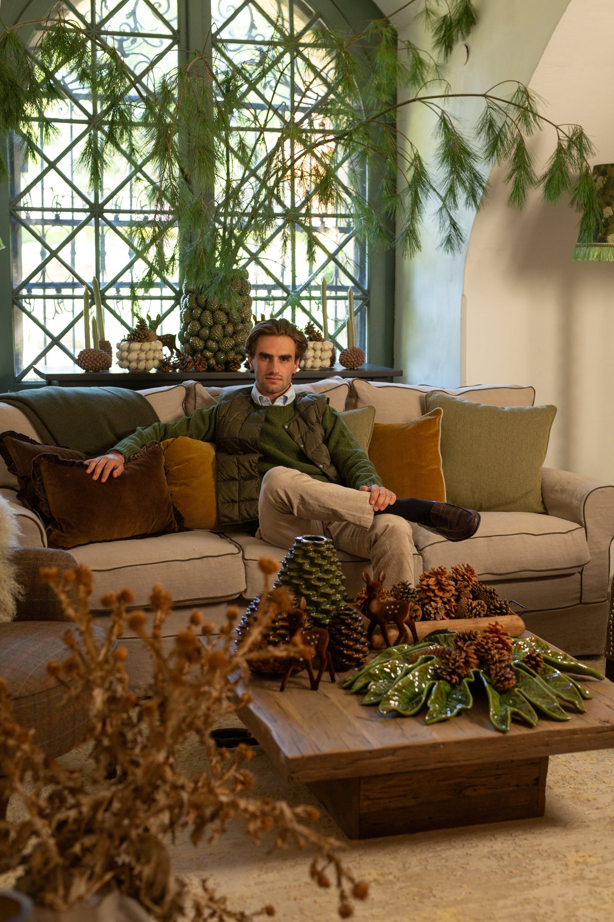 Man on a sofa with green cushions, surrounded by pine branches, cones and winter decorations.