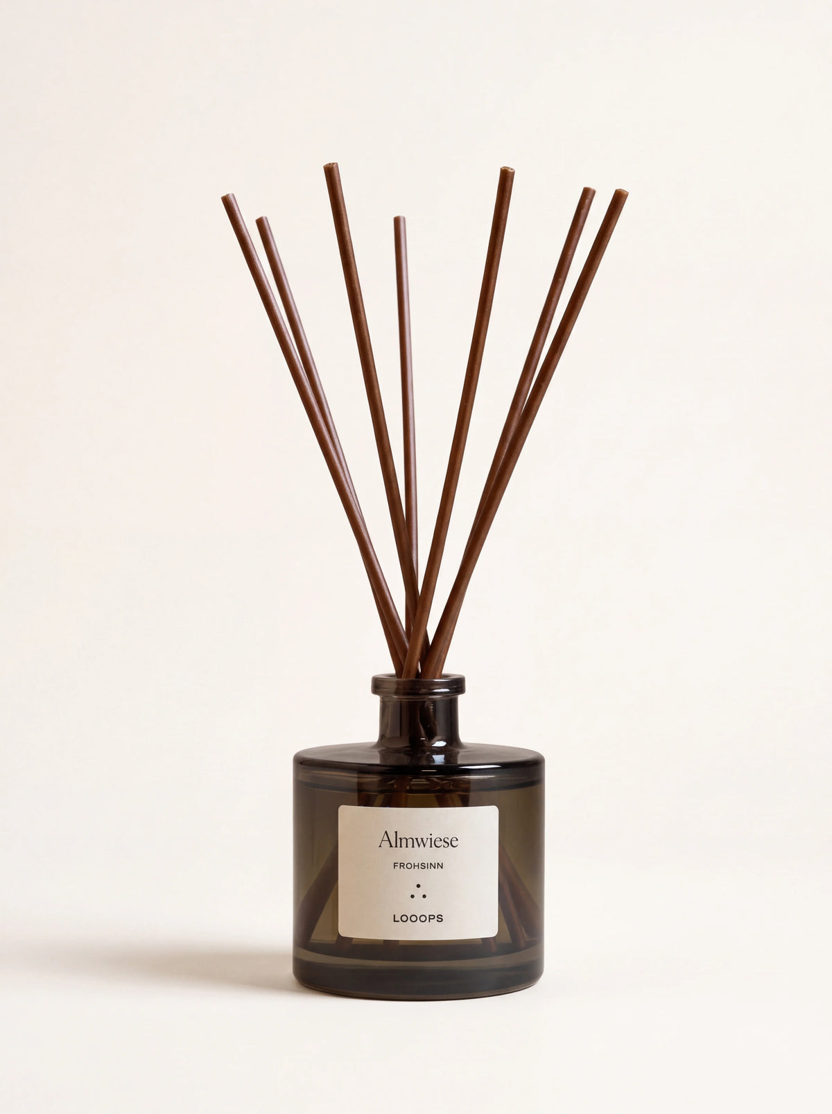 Tinted glass reed diffuser with brown paper reeds and label reading Almwiese FROHSINN LOOOPS