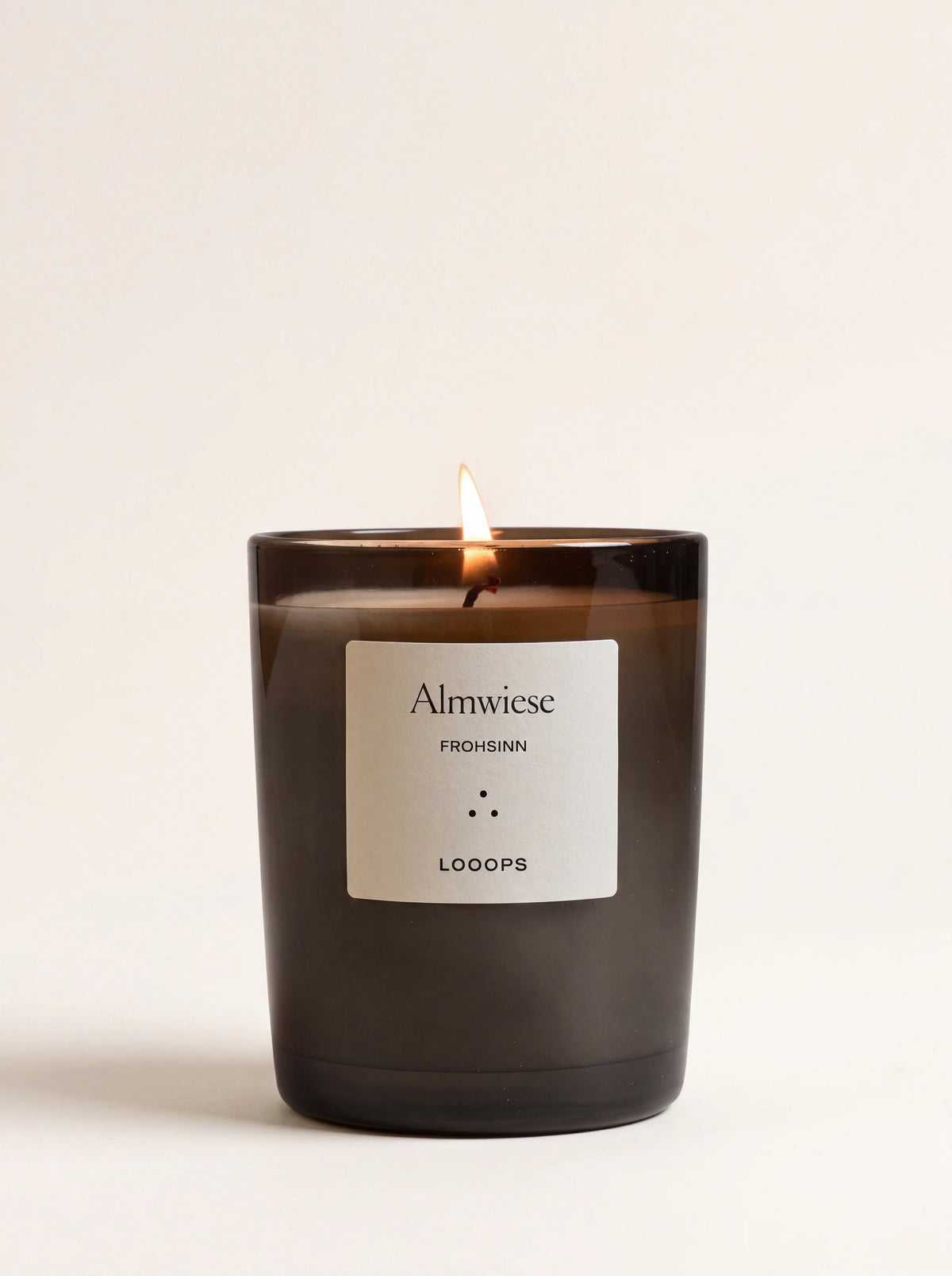 Lit scented candle in a dark glass jar with a white label reading Almwiese Frohsinn Looops
