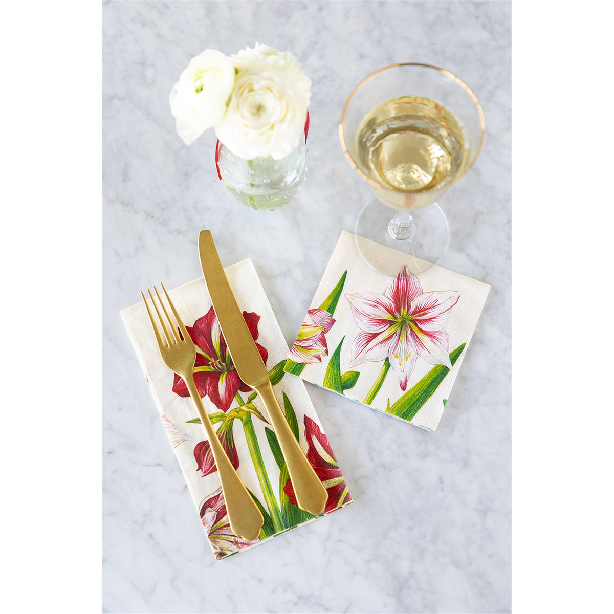 Folded amaryllis paper napkin laid beneath gold cutlery beside a drink and flowers on a tabletop