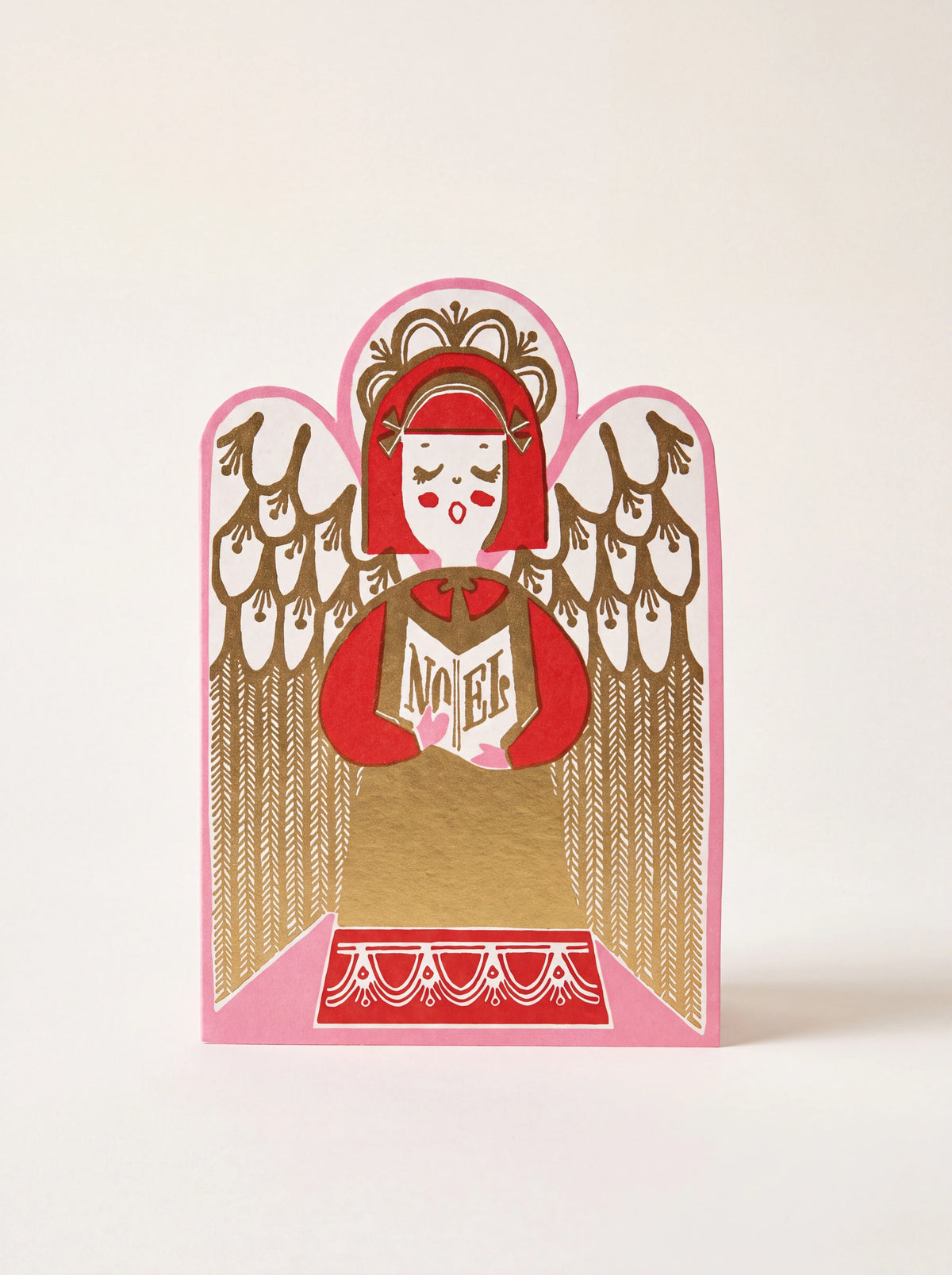 Die-cut paper card shaped as a singing angel in red and gold foil, holding an open book reading NOEL
