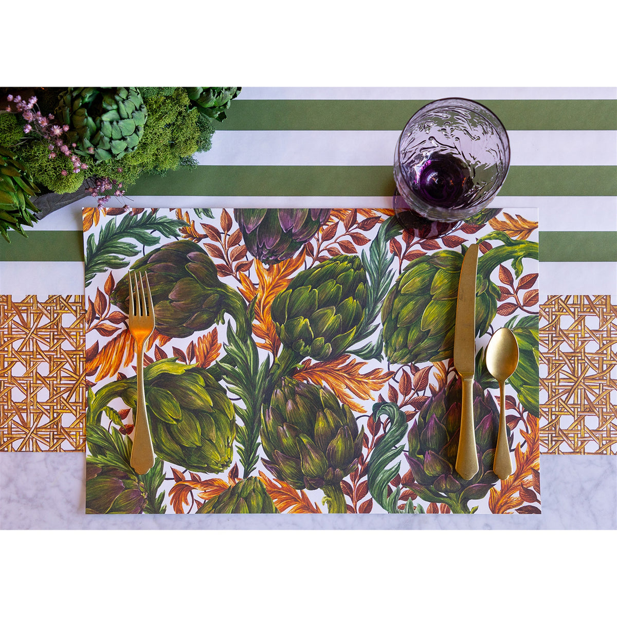 Single artichoke paper placemat set with gold cutlery and a purple glass on a green-striped runner