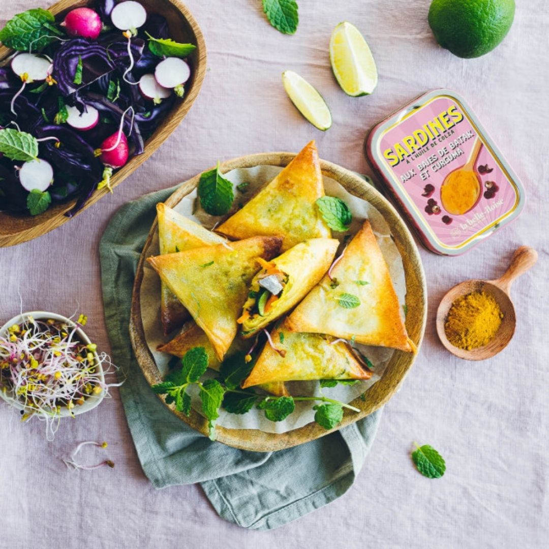 Sardines used as filling in crisp pastries, served on a platter with fresh salad, lime and ground spice