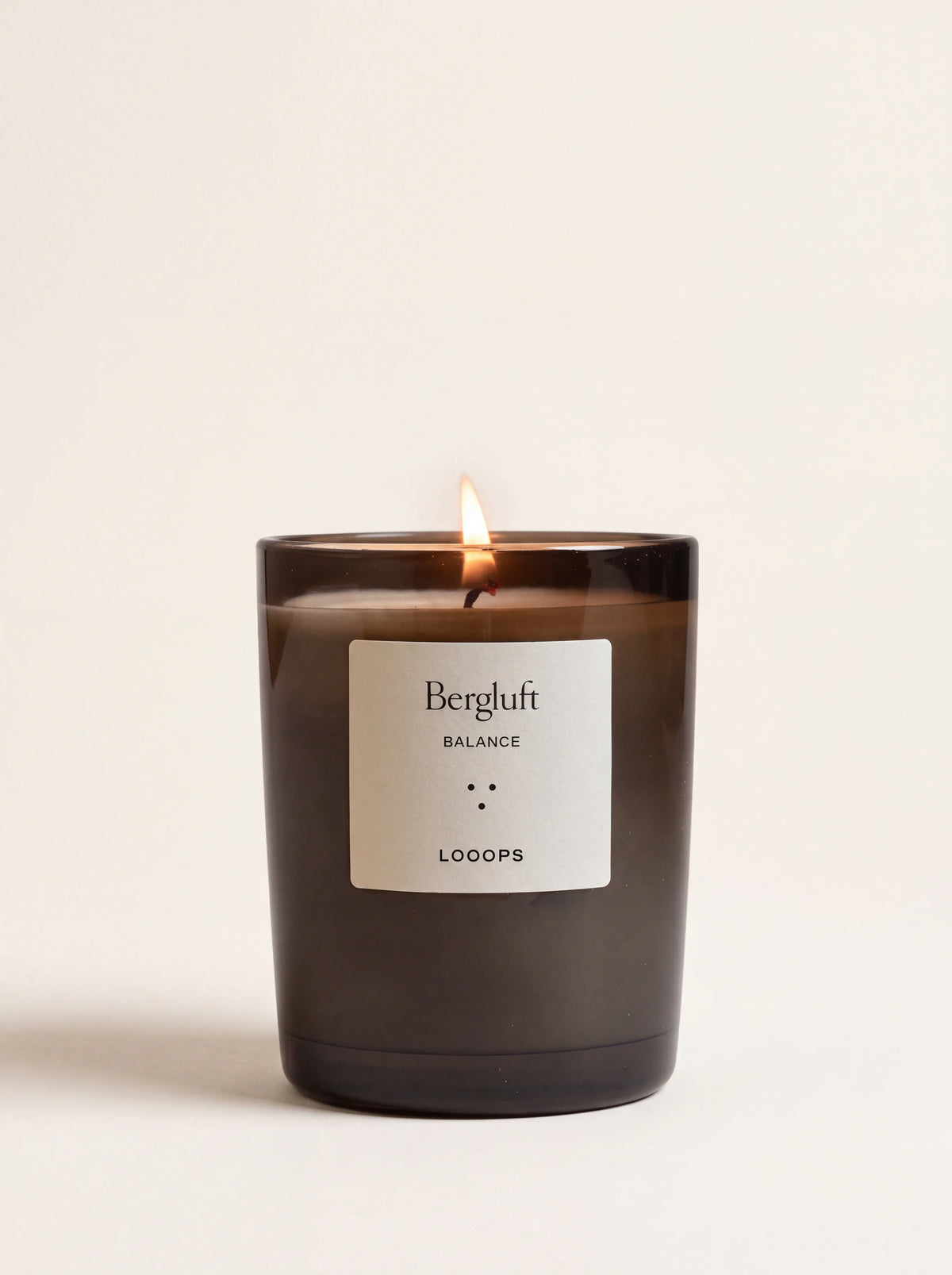 Lit scented candle in a dark tinted glass jar with a label reading Bergluft BALANCE LOOOPS