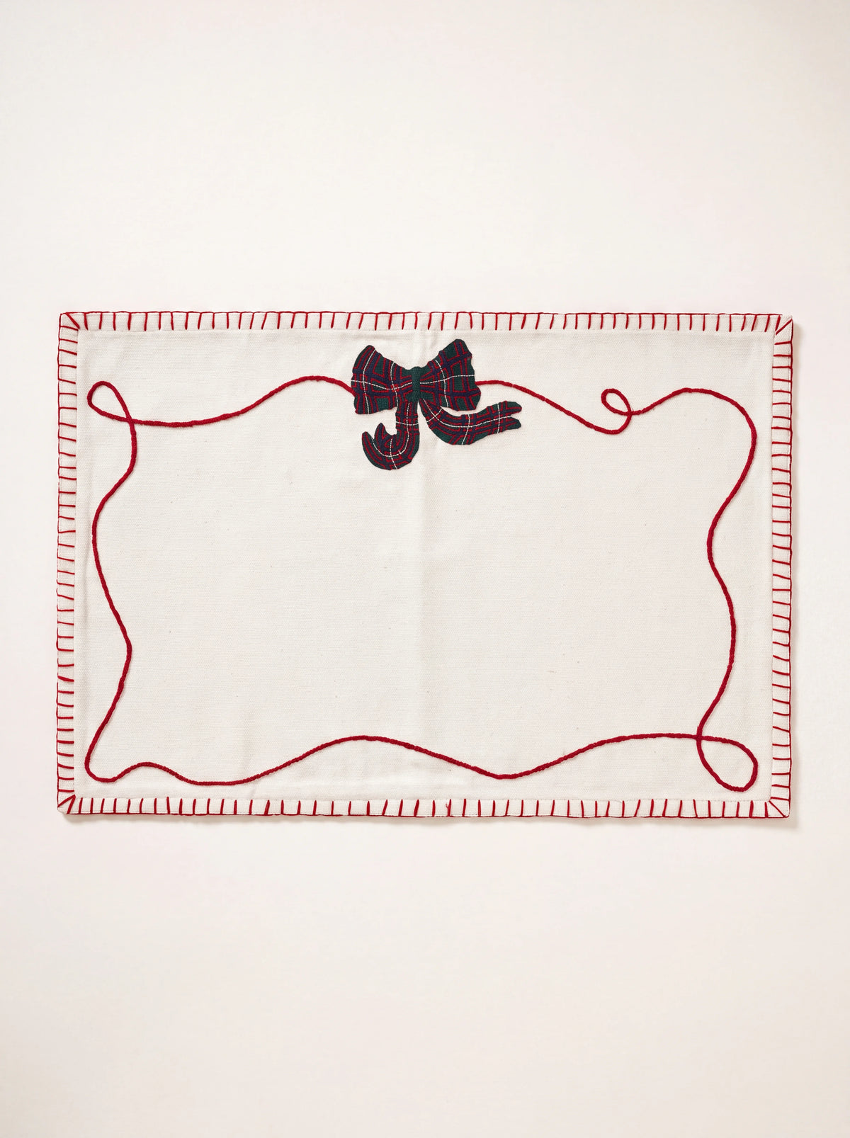 Off-white cotton placemat with red blanket-stitched edges, a green tartan bow, and red ribbon embroidery