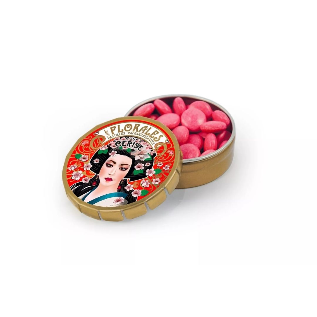 Open round metal tin filled with small pink cherry pastilles, with the illustrated lid resting in front