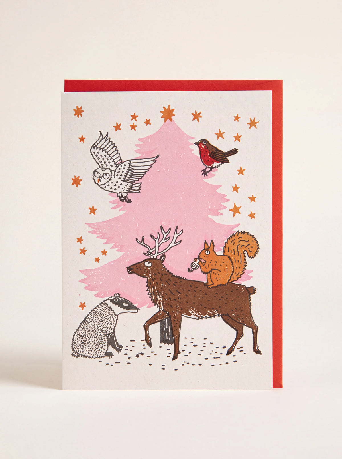 Paper Christmas card illustrated with woodland animals around a pink tree, paired with a red envelope