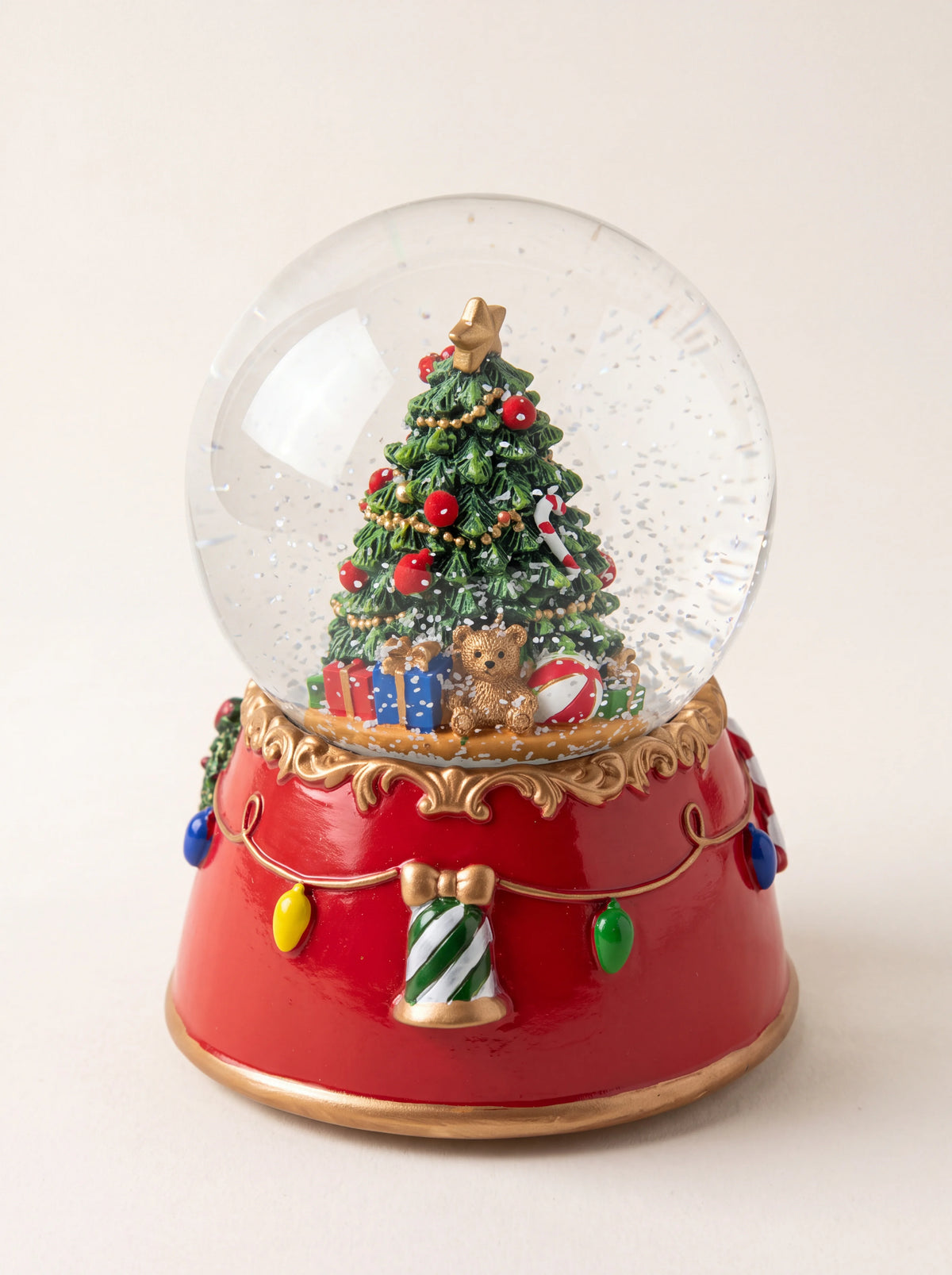 Christmas tree snow globe with gifts and a teddy bear inside, on a red base with gold trim and light garlands