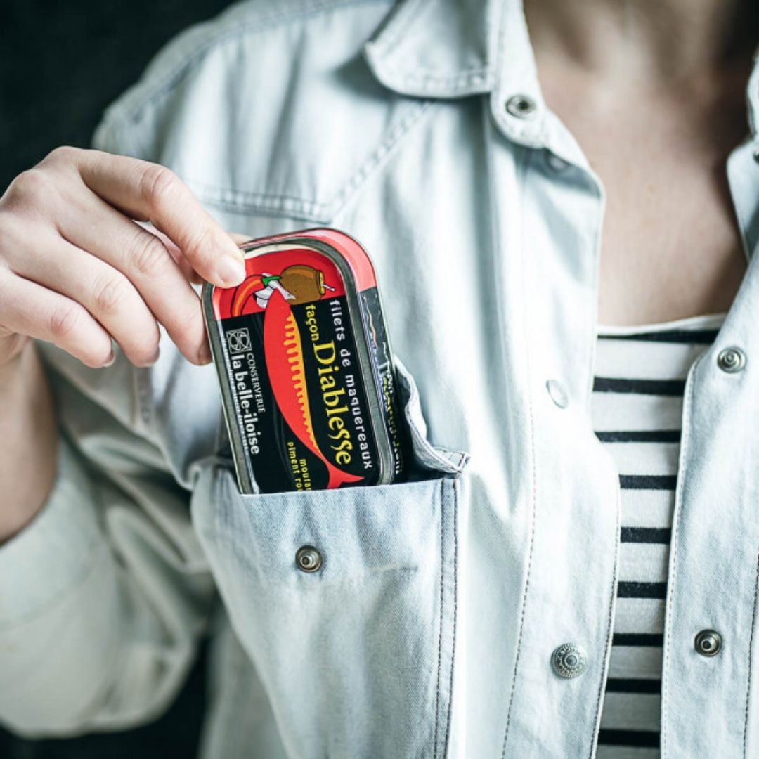 Tinned mackerel fillets held in the breast pocket of a light denim shirt, showing scale