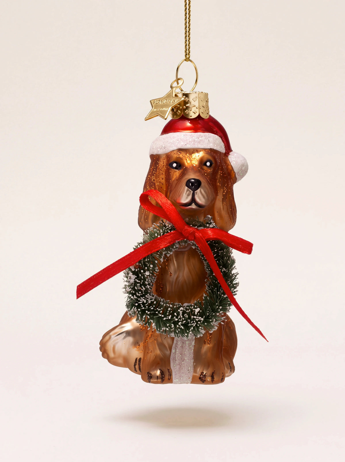 Glass dog Christmas ornament wearing a red Santa hat and holding a frosted wreath with a red bow