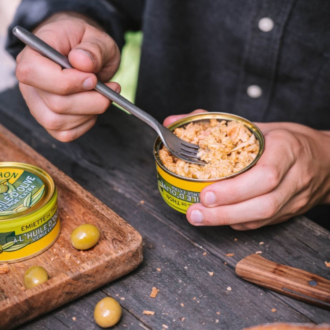 Open tin held in hands, with a fork showing the pale, coarsely flaked tuna meat in olive oil
