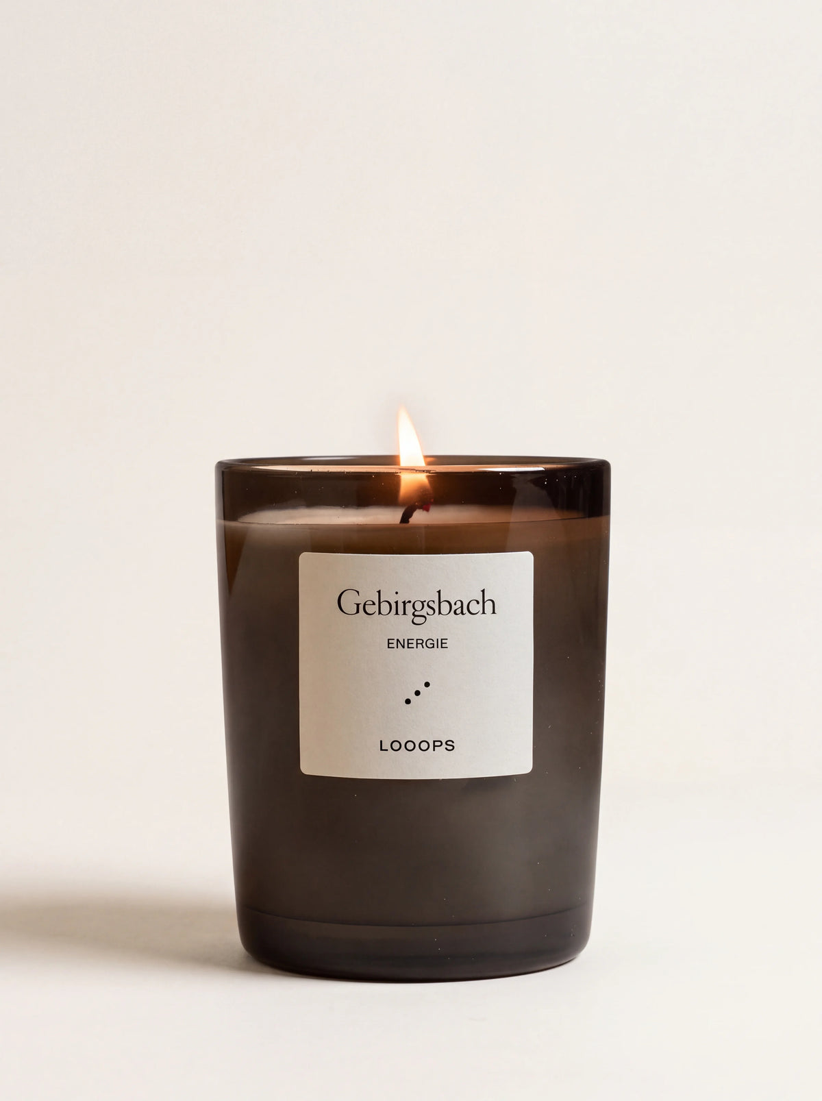Lit scented candle in a dark smoked glass jar with a label reading Gebirgsbach