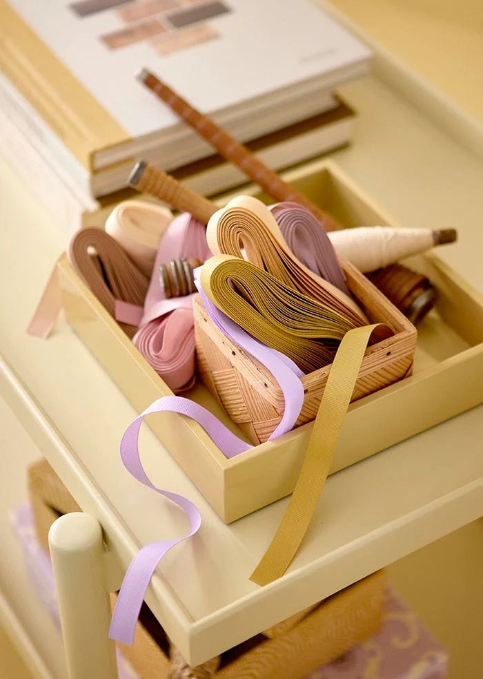 Grosgrain ribbons stored in small craft boxes, showing an unrolled lilac ribbon trailing over a tray edge