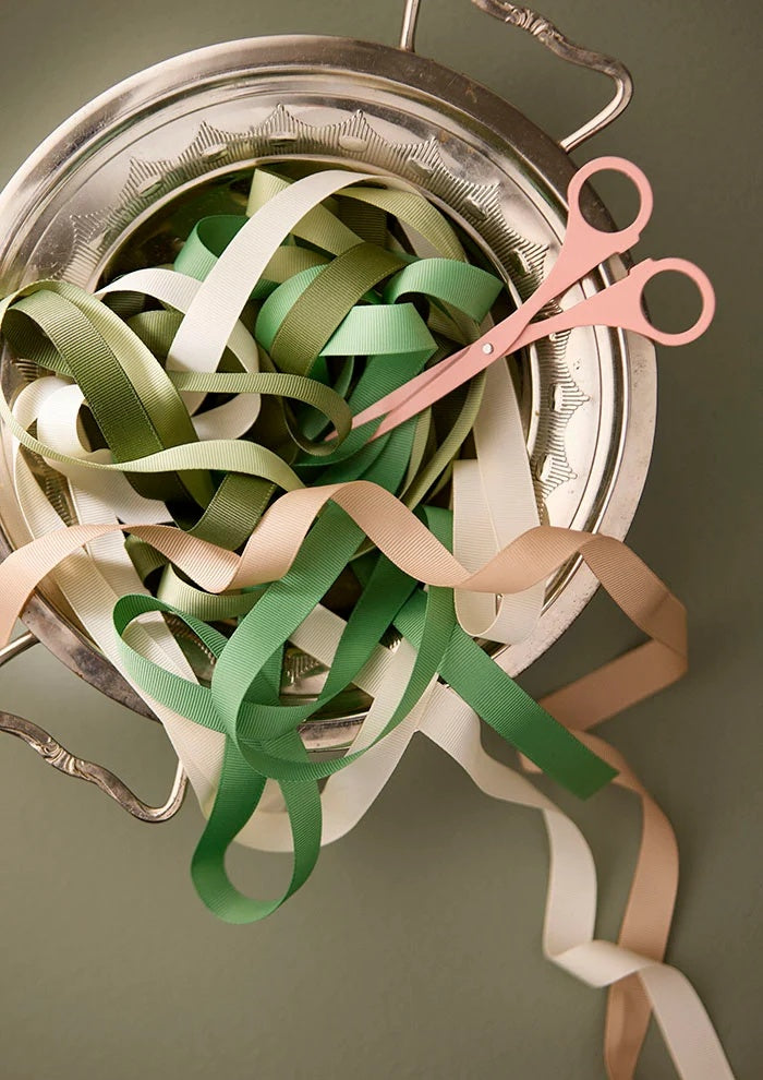 Lengths of unrolled grosgrain ribbon loosely gathered inside a silver metal dish beside pink craft scissors