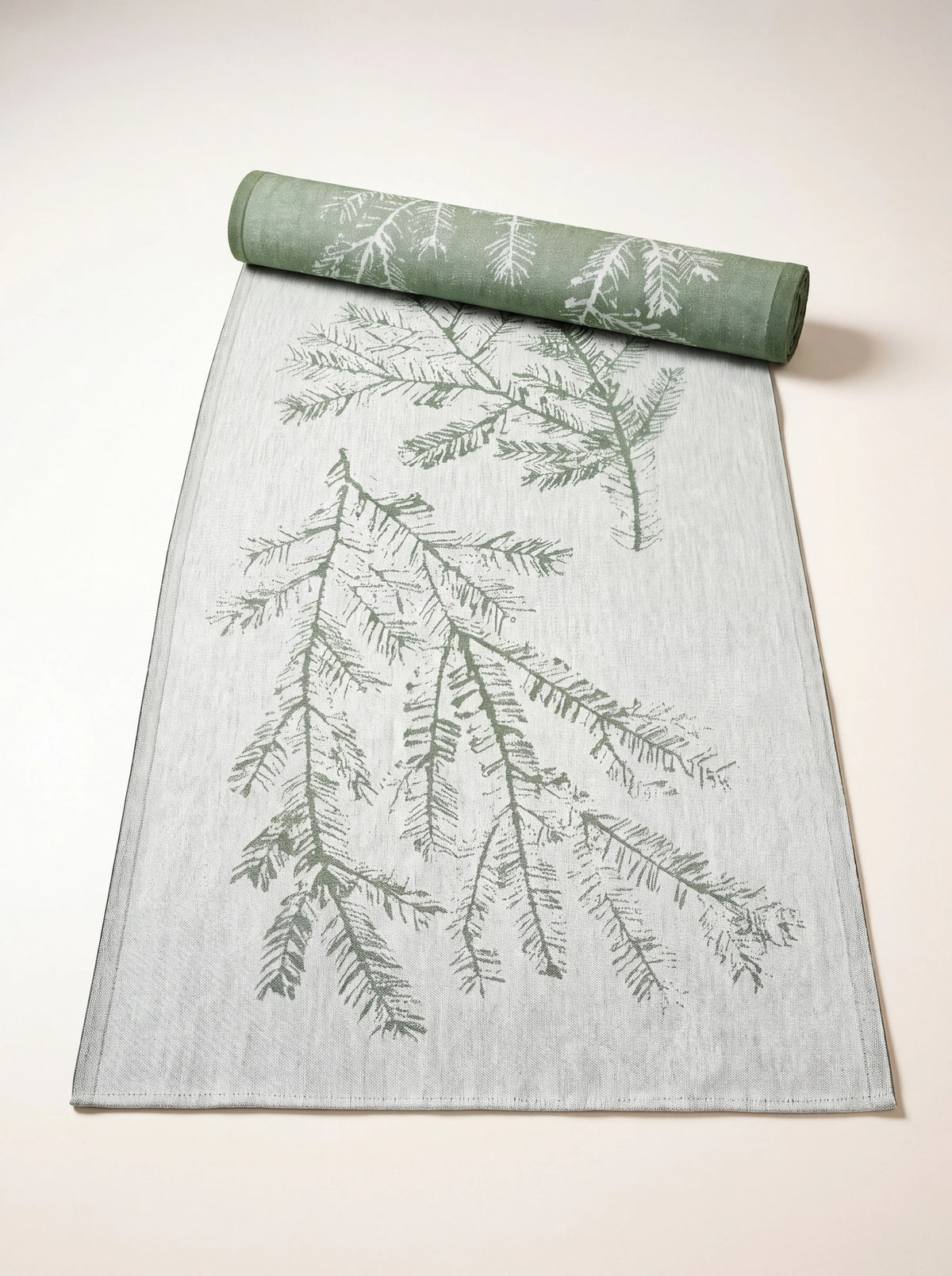 Linen and cotton table runner partially rolled, showing a green pine branch design on a light woven ground