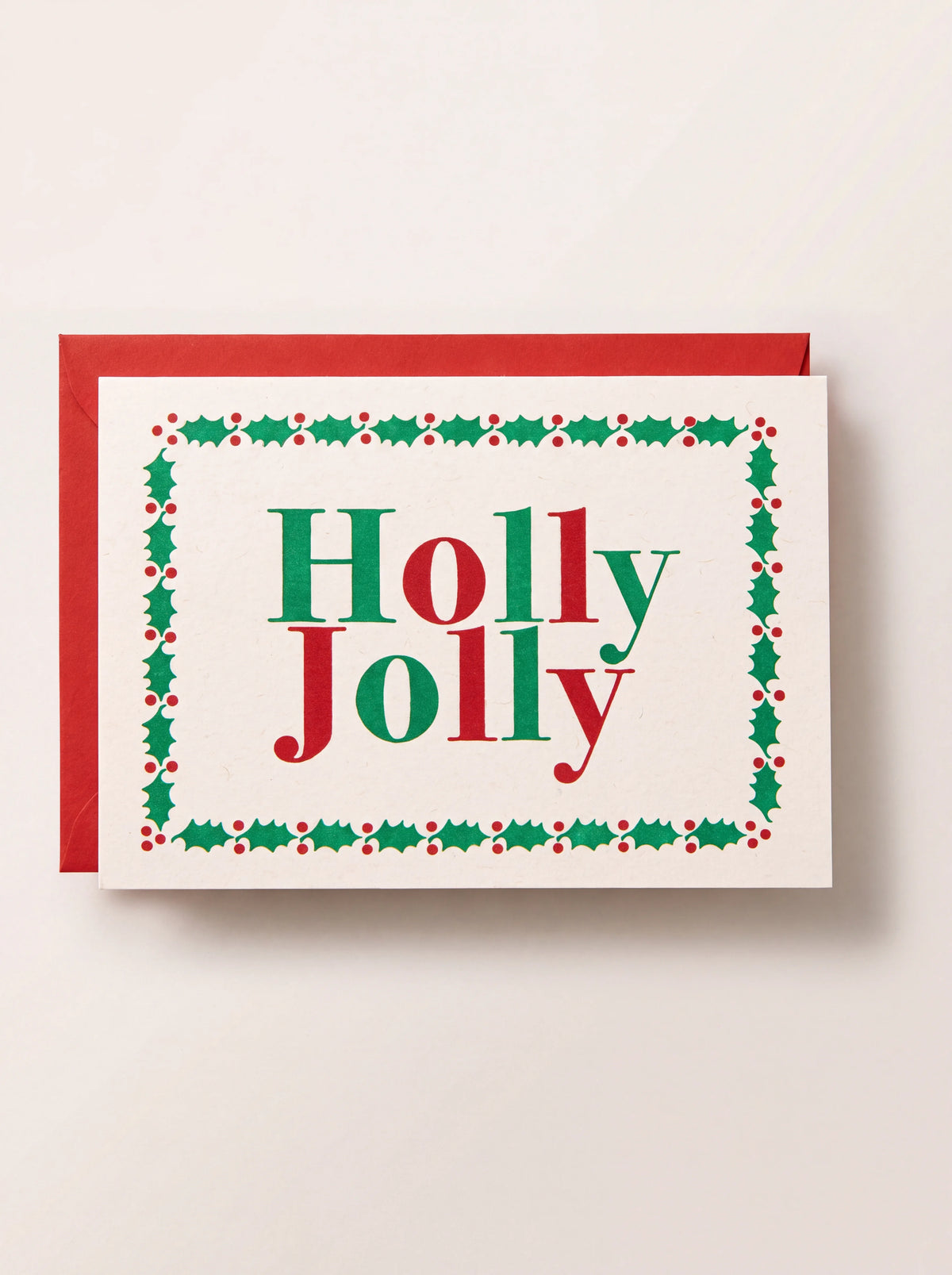 Paper Christmas card reading Holly Jolly with a holly leaf border, shown against a red envelope