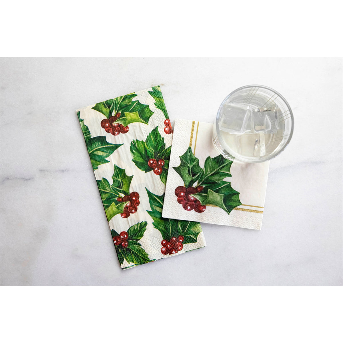 Folded holly paper guest napkin shown beside a square cocktail napkin and a water glass