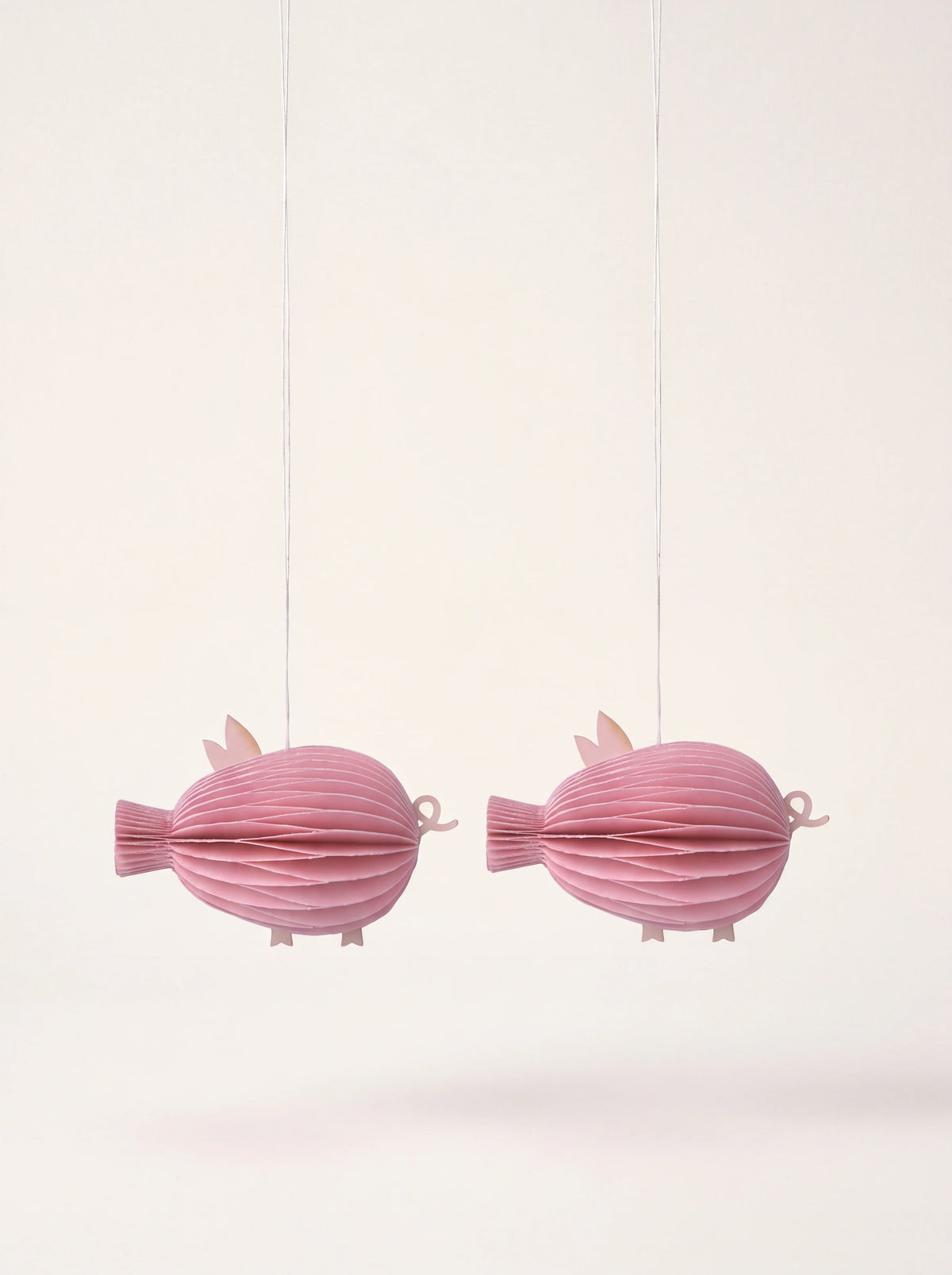 Pink honeycomb paper pig ornaments suspended by fine hanging threads, shown as a set of two