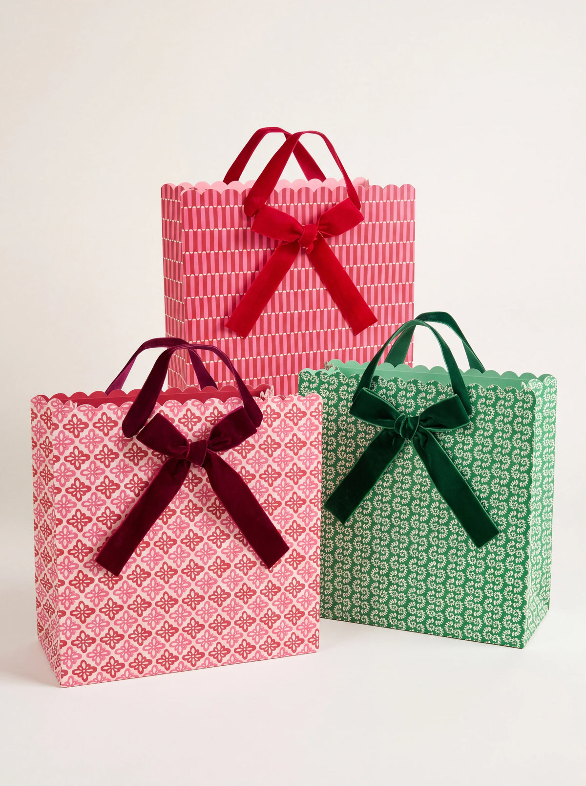 Set of three patterned paper gift bags in pink, red and green with scalloped rims and velvet ribbon bows