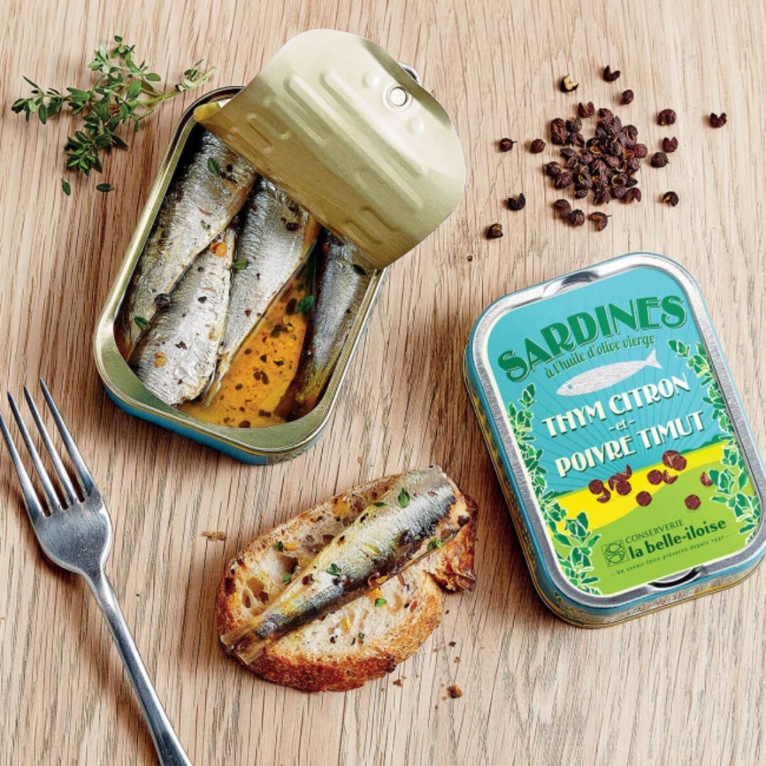 Open tin showing sardines in seasoned oil, with one sardine served on toasted bread