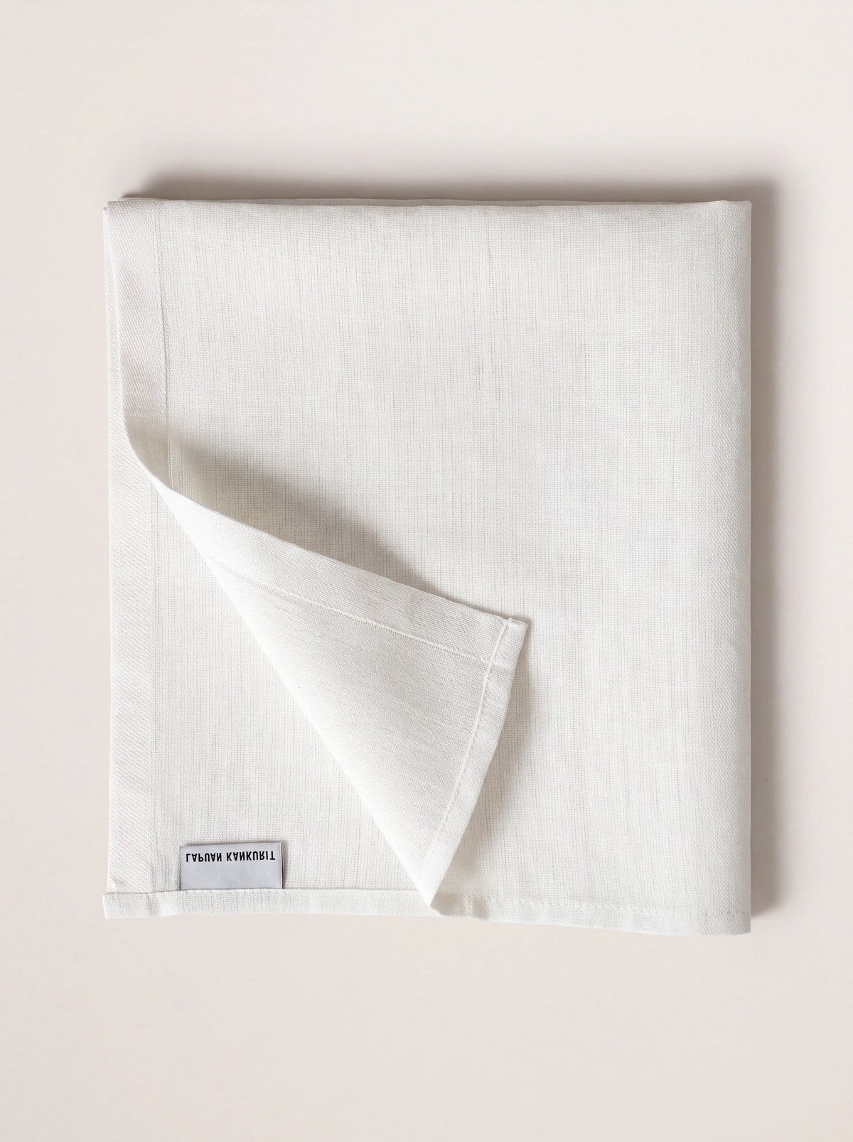 Square white fabric napkin folded with one corner turned up to show the stitched hem and label