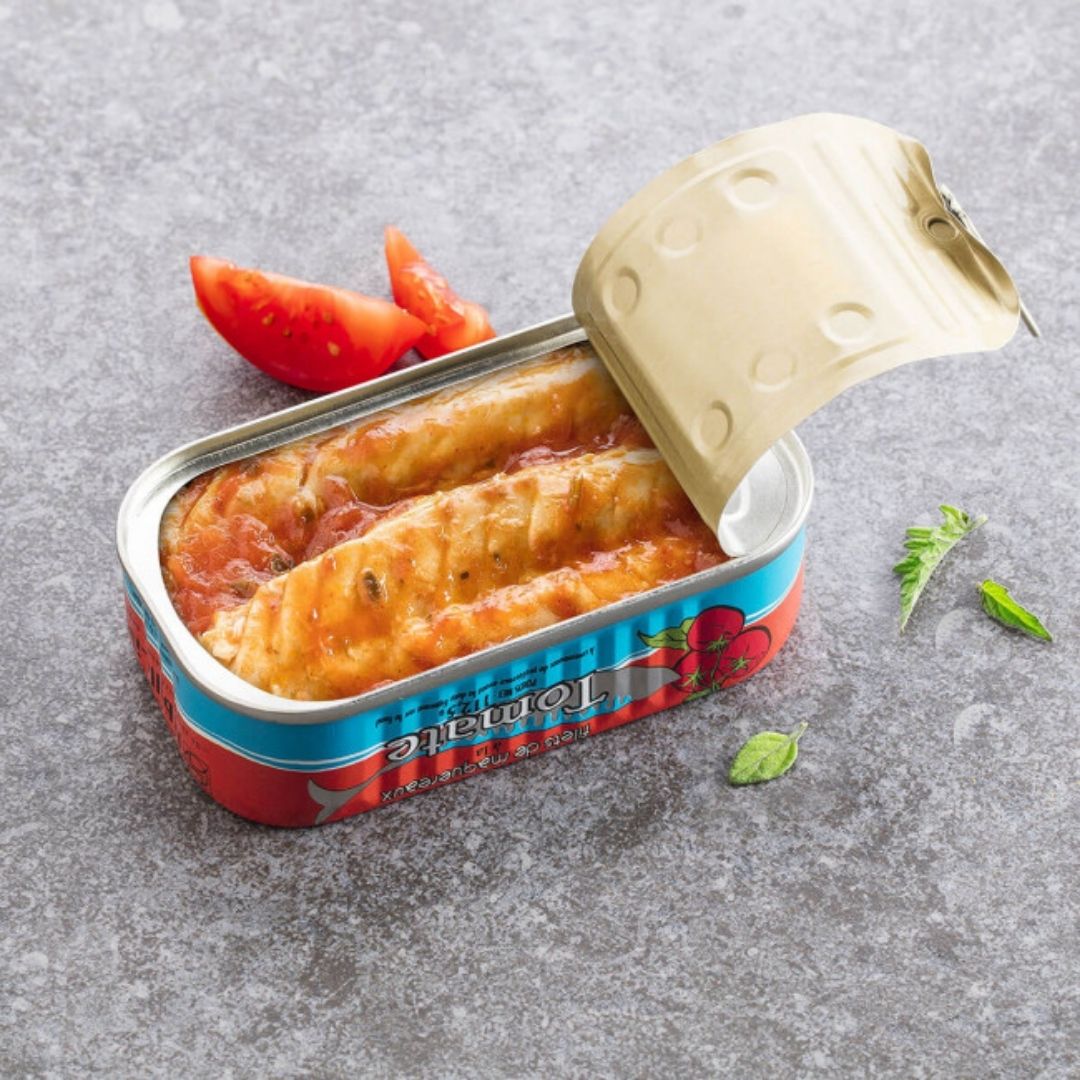 Open tin showing mackerel fillets coated in tomato sauce, with fresh tomato slices beside it