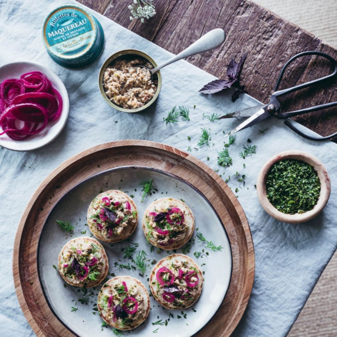Mackerel rillettes served from the open tin onto blinis garnished with pickled red onion and herbs