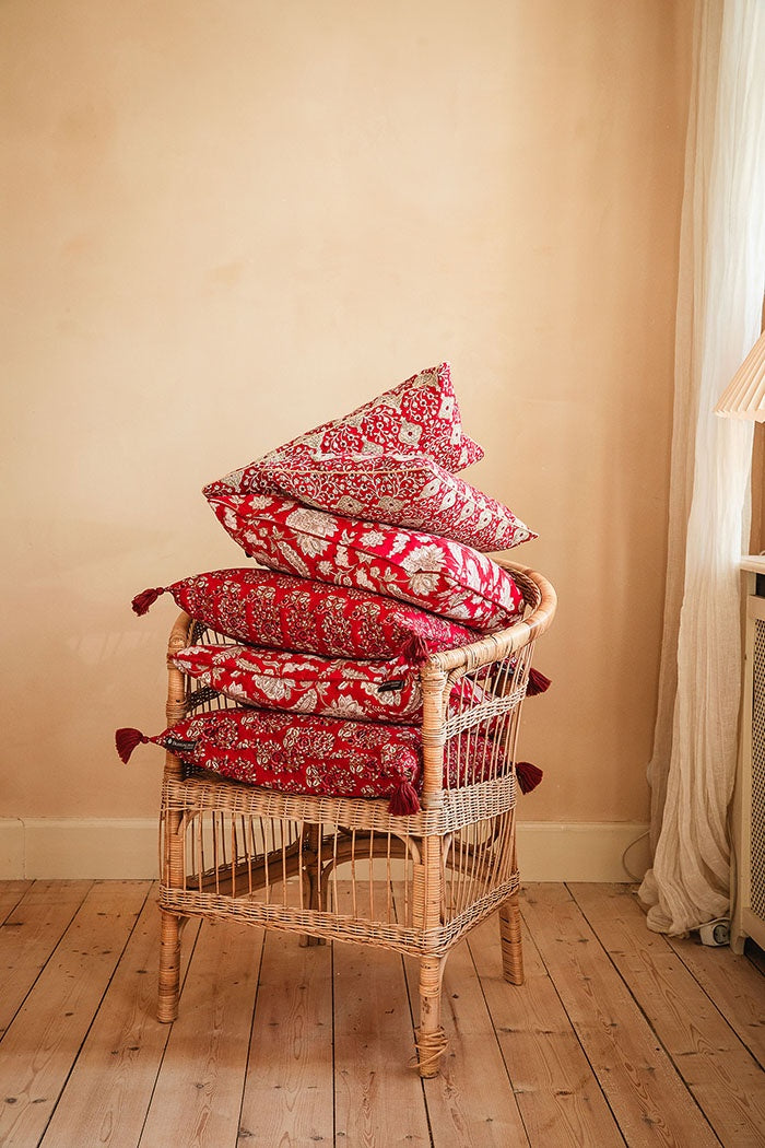 Stack of red patterned cotton cushions with tassels piled high on a woven wicker armchair