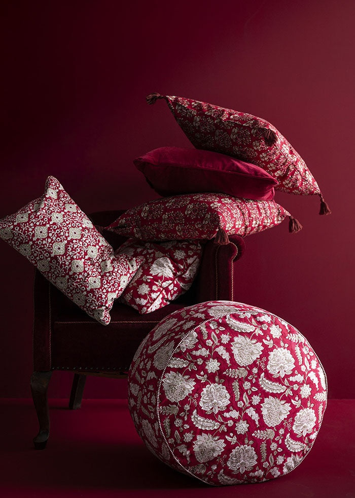 Ruby floral cushions arranged on a dark red armchair alongside a matching round floor pouffe