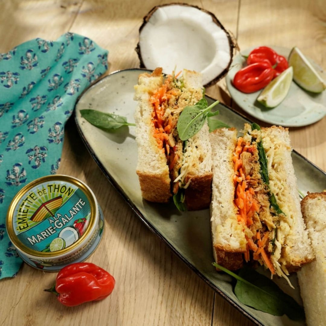 Flaked tuna sandwich cut in half with shredded carrot and greens, on a platter beside the tin