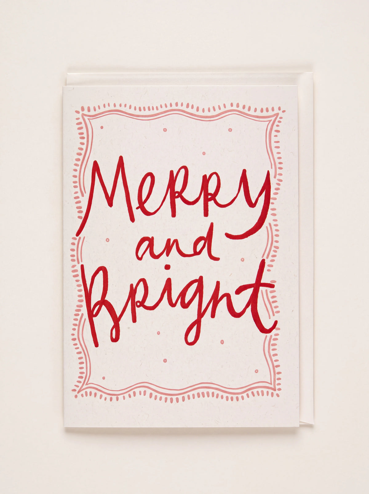 Letterpress greeting card with red Merry and Bright lettering inside a pink border, shown with its white envelope