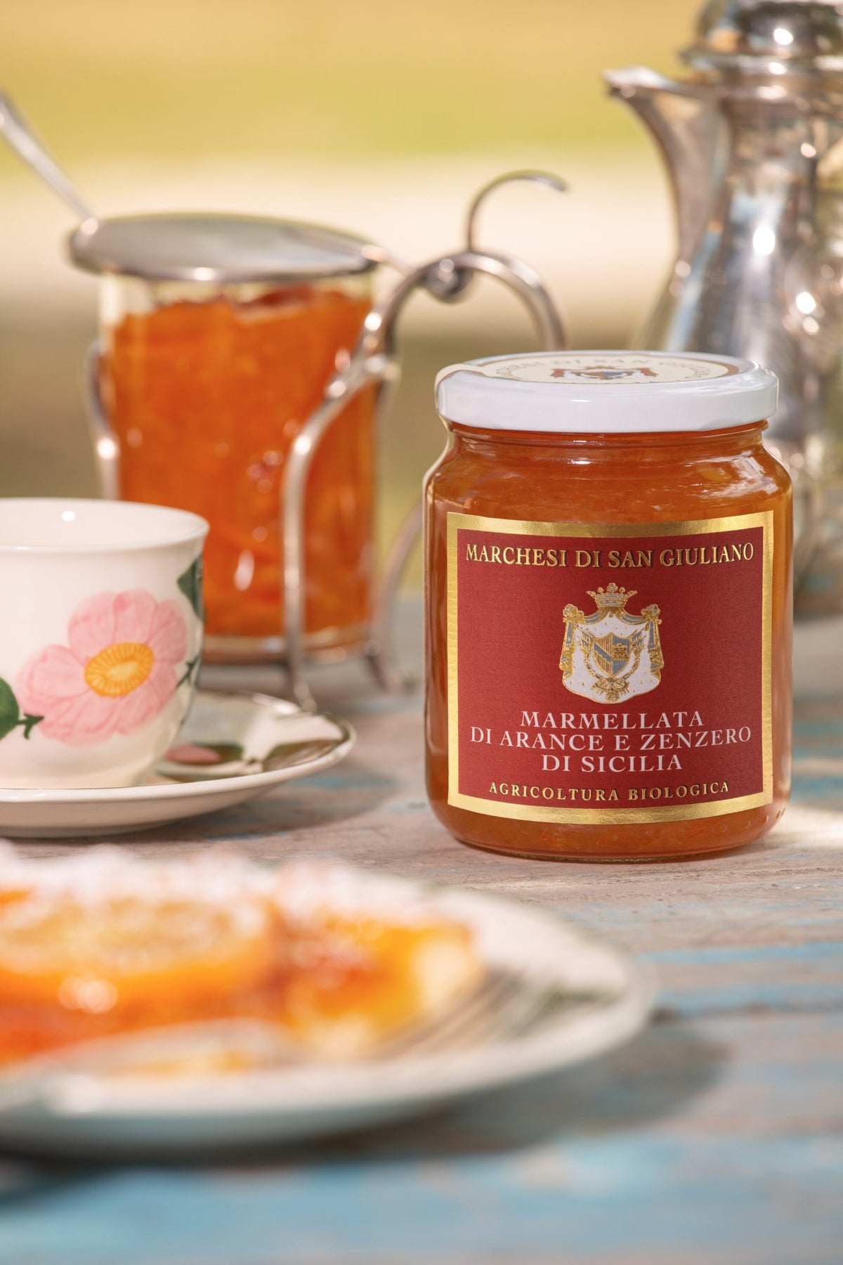 Orange and ginger marmalade jar set on a table with a floral teacup, tart and silver pot