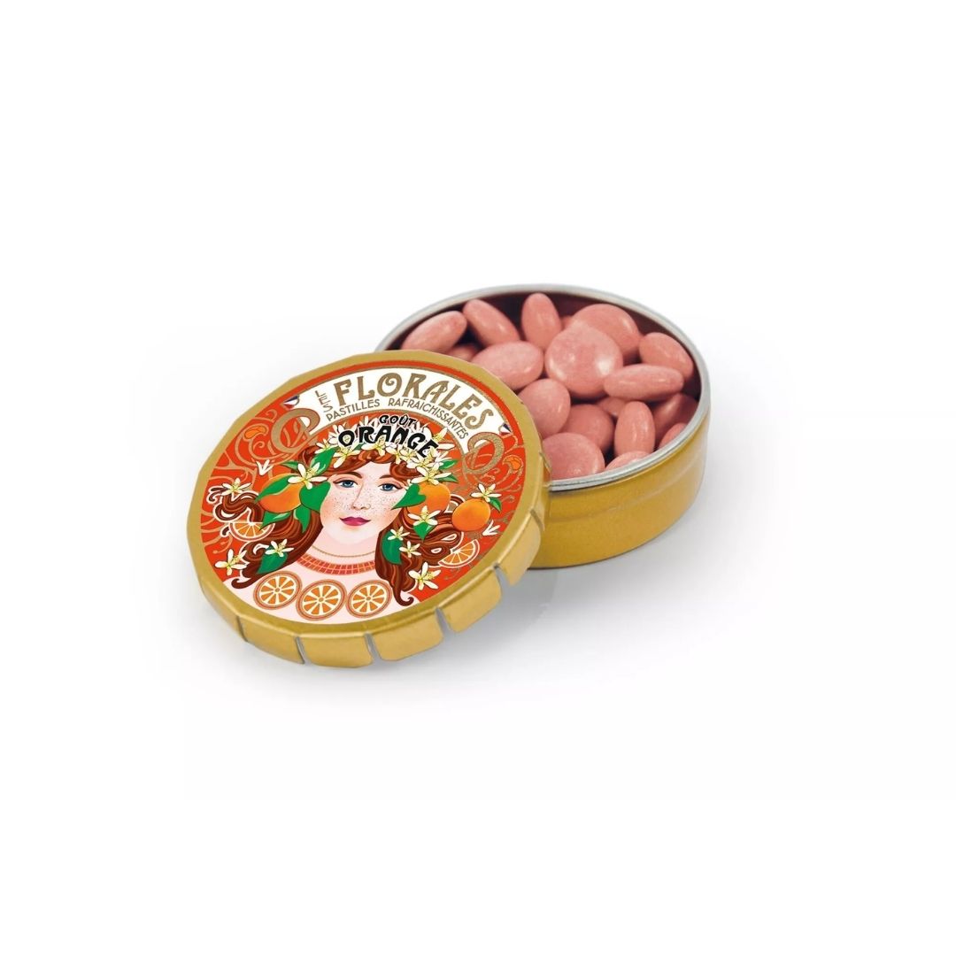 Open gold-coloured tin filled with small round pastilles, with the illustrated lid resting in front
