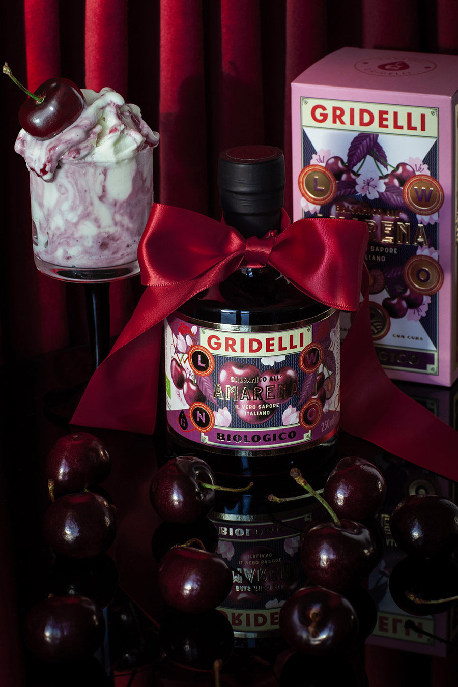 Bottle tied with a red satin ribbon, shown beside its pink gift box, fresh cherries and a cherry dessert
