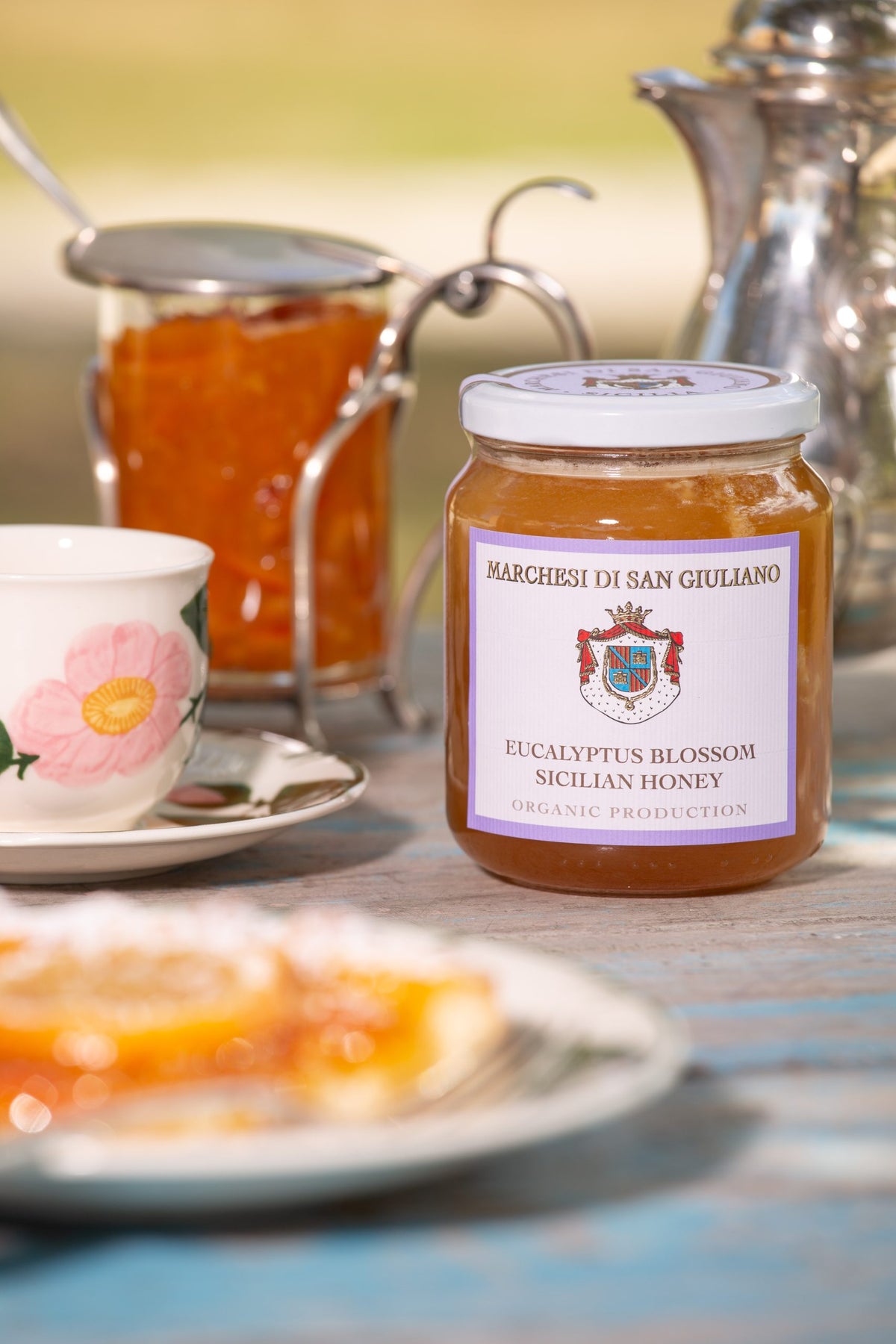 Jar of eucalyptus honey on a table alongside a floral teacup, a silver pot and a citrus pastry