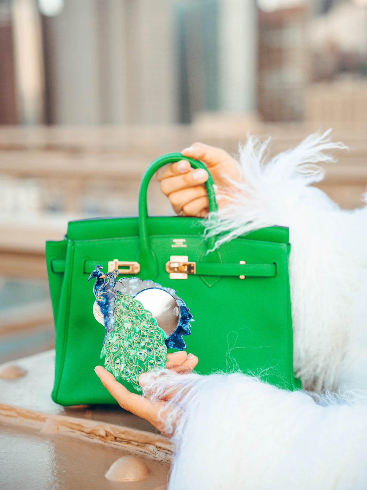 Peacock compact mirror held open in one hand, showing scale next to a green handbag