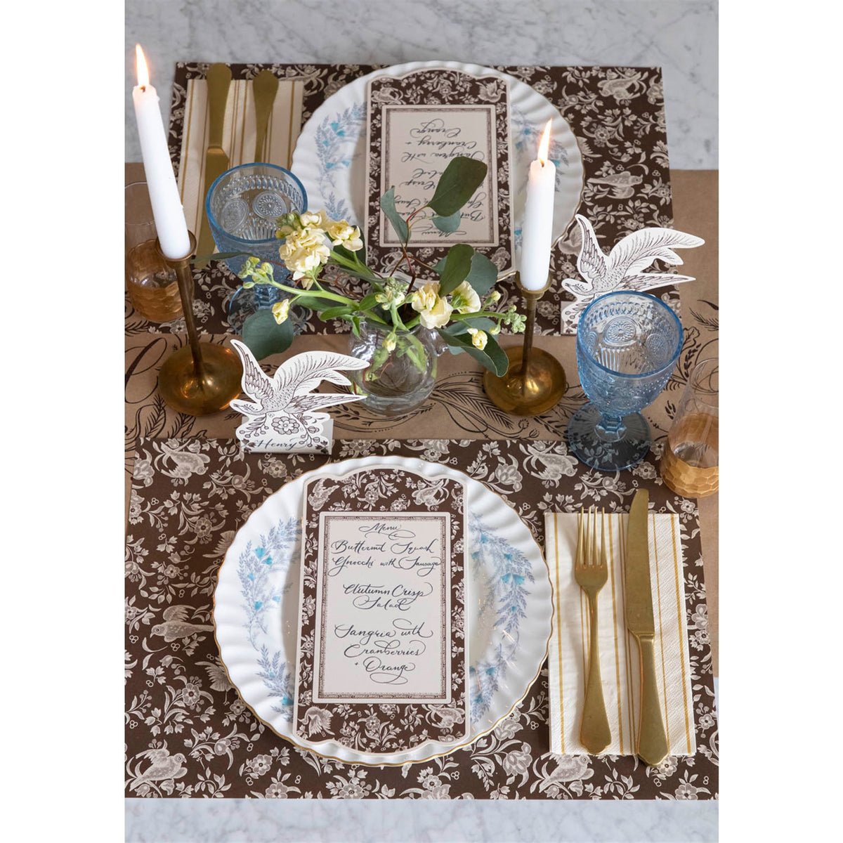 Brown peacock paper placemats arranged in a dining table setting with plates, cutlery and candles