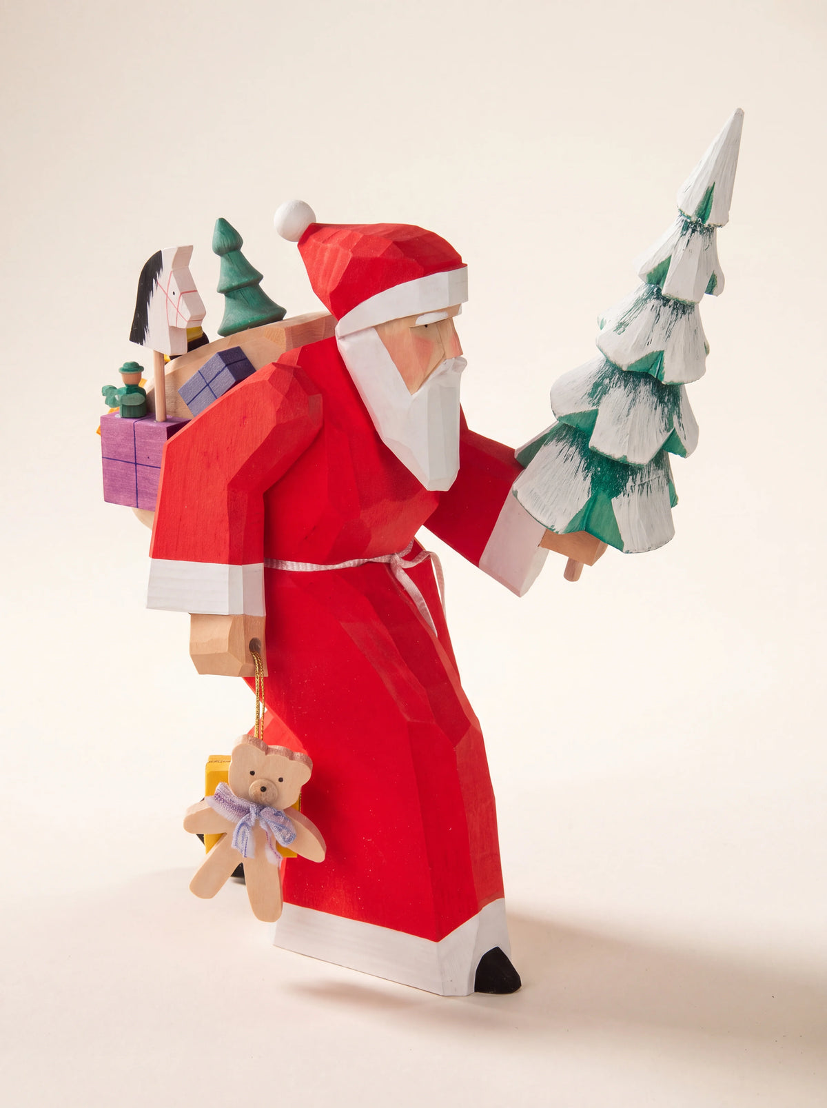Carved wooden Santa Claus figure in a red robe carrying a snow-tipped tree, a sack of toys and a teddy bear