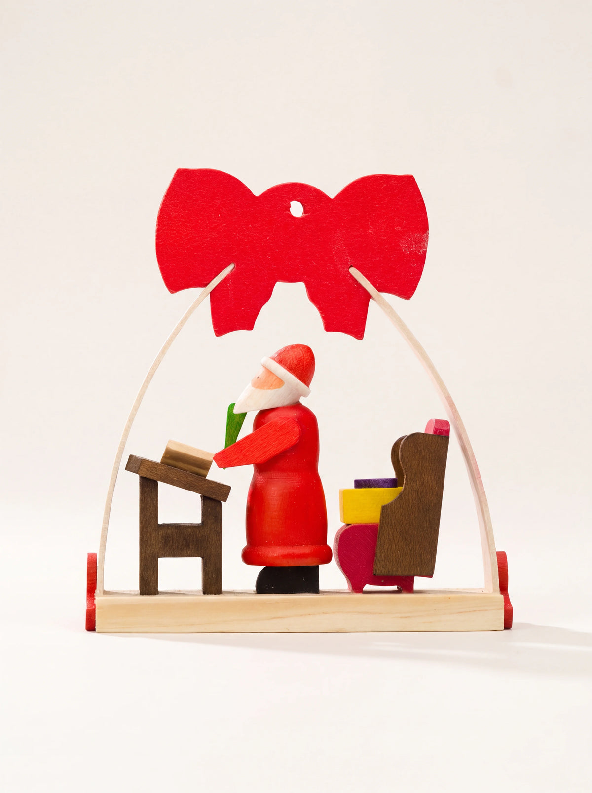 Wooden Santa ornament with an arched frame and red bow, showing Santa at a desk beside an armchair