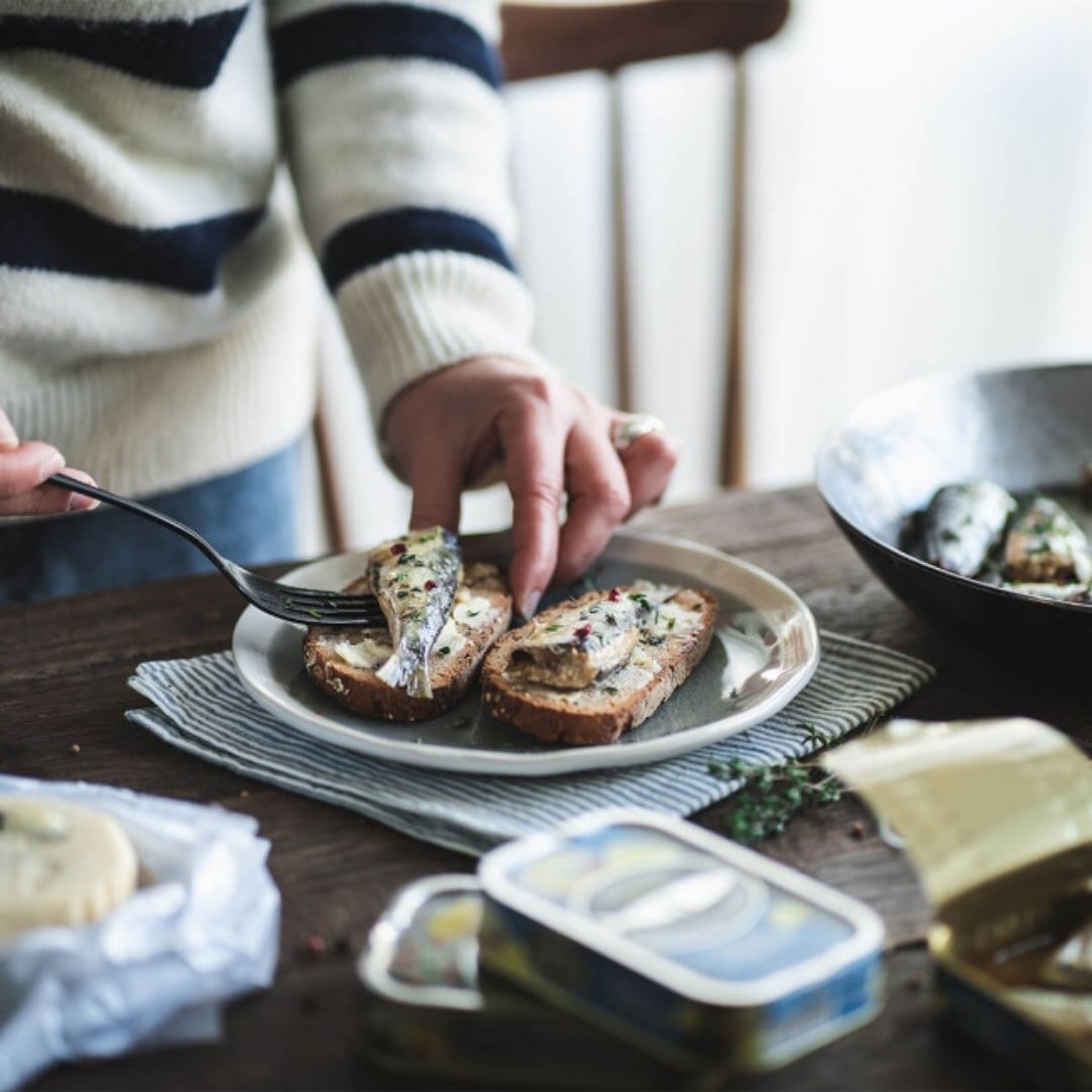 Placing a cooked sardine with a fork onto crusty bread on a plate, next to an open tin