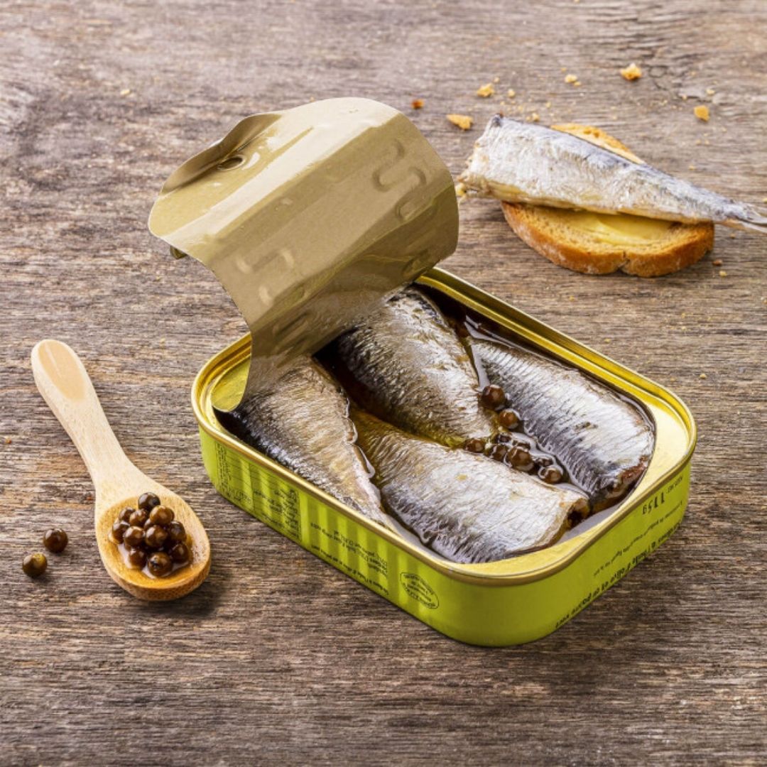 Opened tin displaying sardines packed in olive oil with green peppercorns, beside toast and a wooden spoon