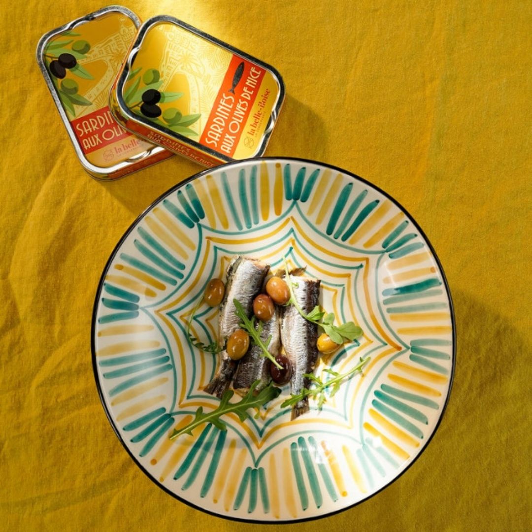 Sardines and whole Niçoise olives garnished with leaves on a patterned plate beside two tins