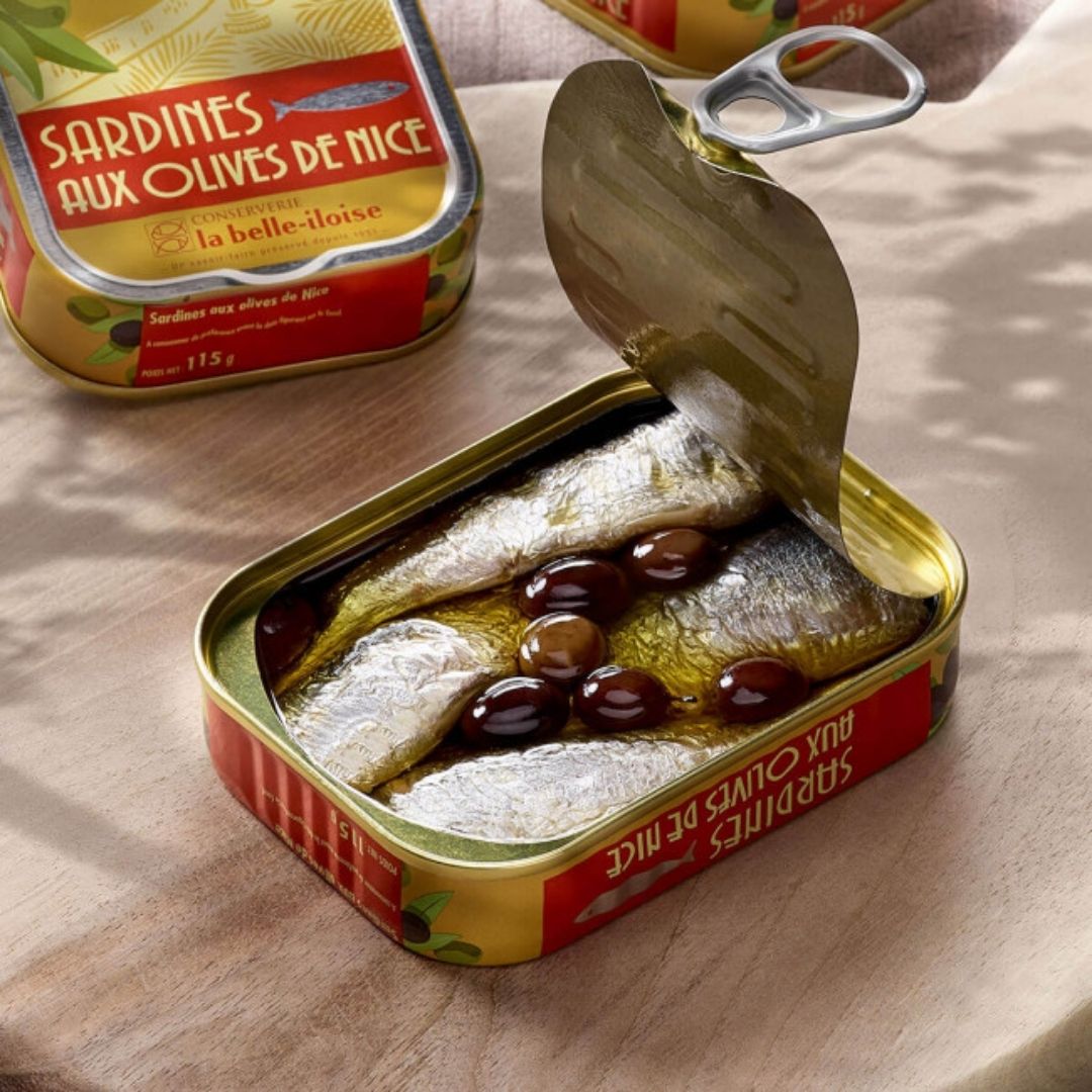 Opened tin of sardines packed in olive oil with dark whole Niçoise olives visible under the lid