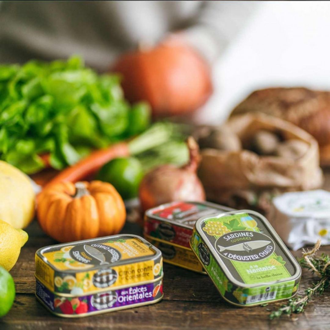Tin of sardines with oriental spices on a wooden table among assorted fresh vegetables and other tins
