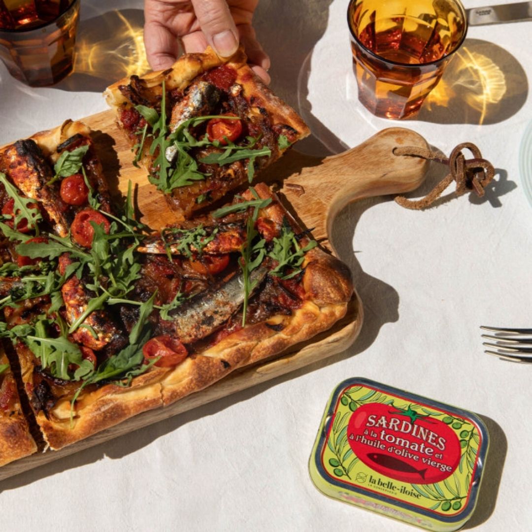 Sardine and tomato tart topped with rocket on a wooden board next to the tin