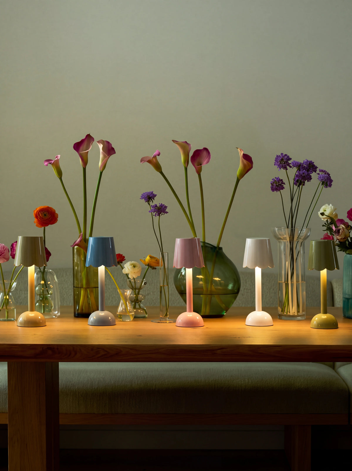Illuminated scalloped table lamps in various colours arranged along a wooden table with flower vases