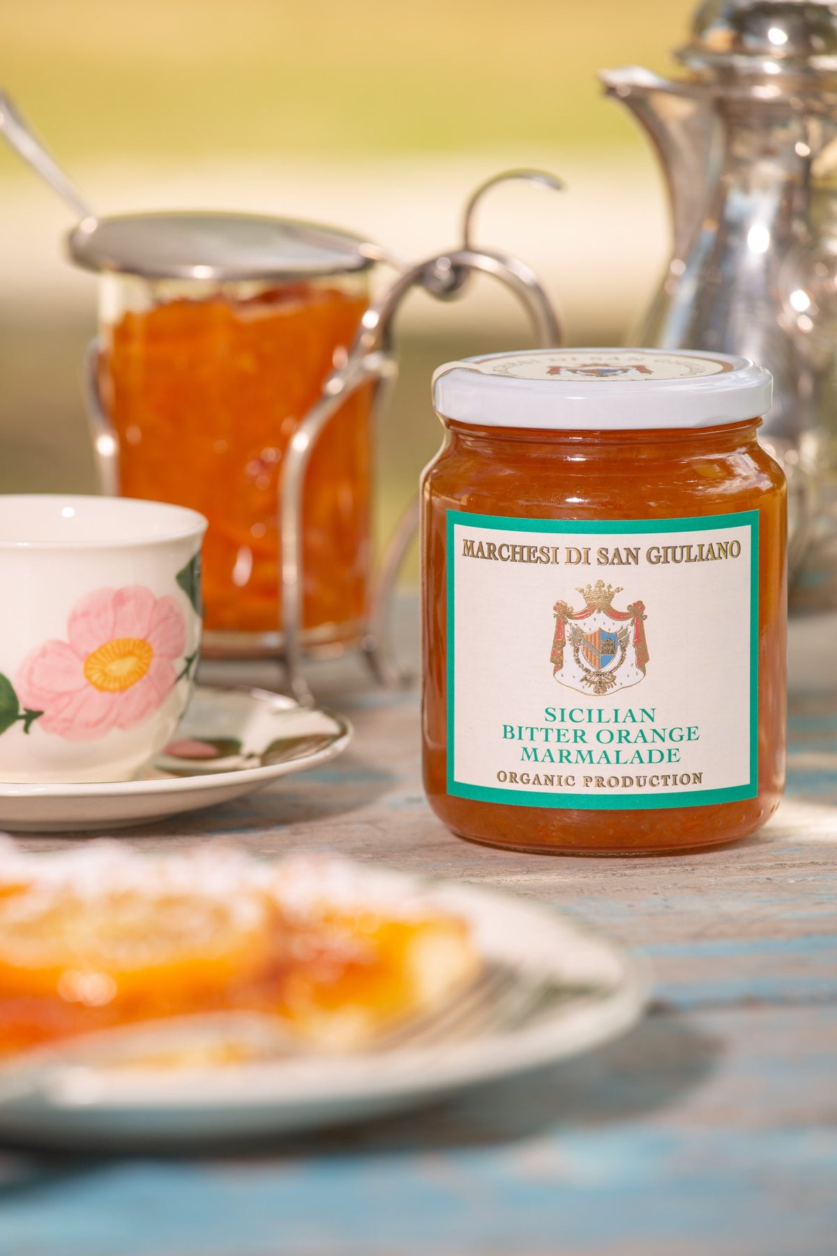 Glass jar of bitter orange marmalade on a table beside a teacup, silver pot and orange pastry