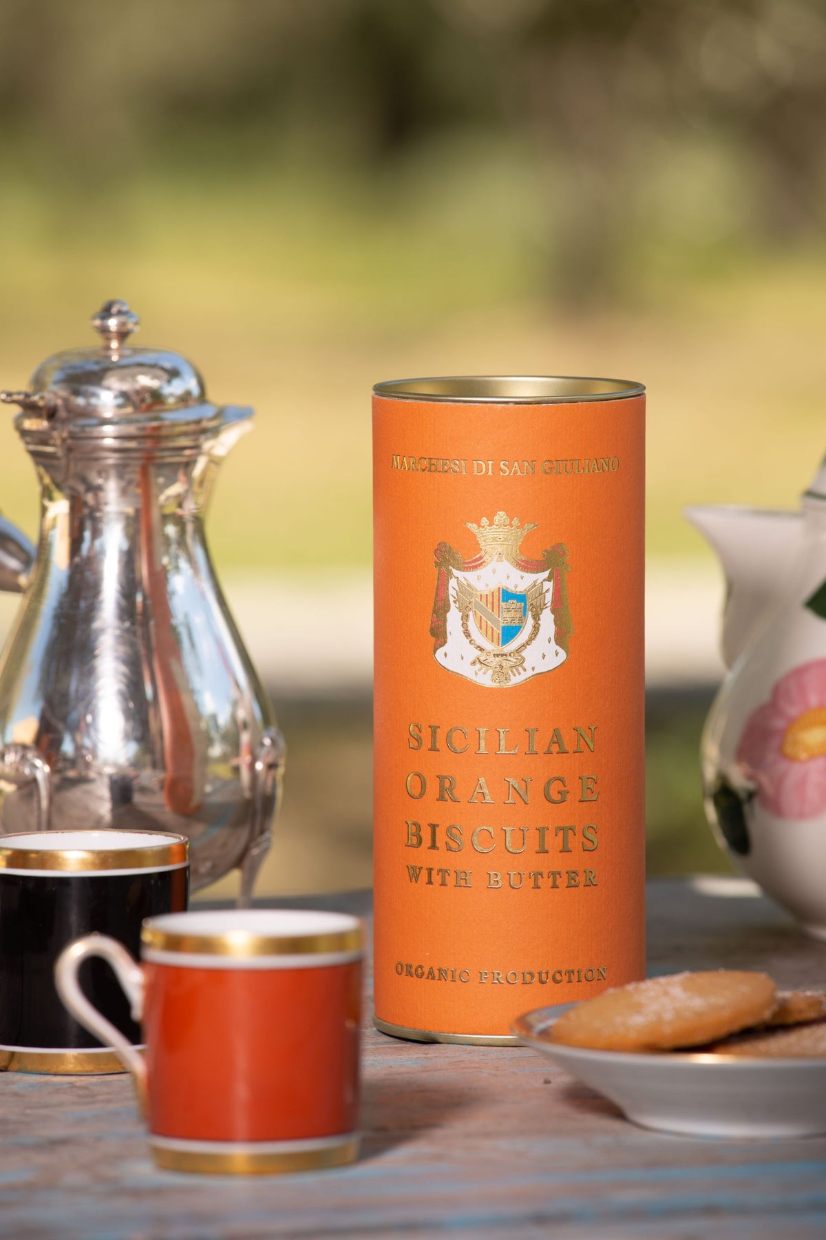 Round Sicilian orange biscuits on a saucer beside the orange tin, small coffee cups and a silver pot